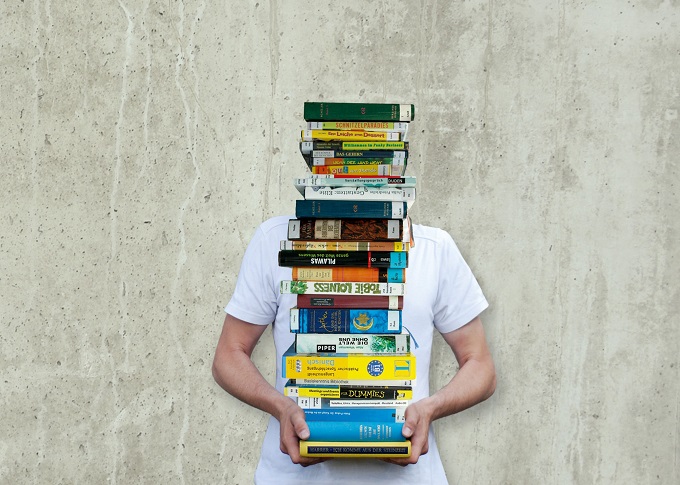 Muskulöser Mann trägt einen riesigen Stapel Bücher. Man sieht nur die Arme, der Stapel überdeckt den Körber und den Kopf.