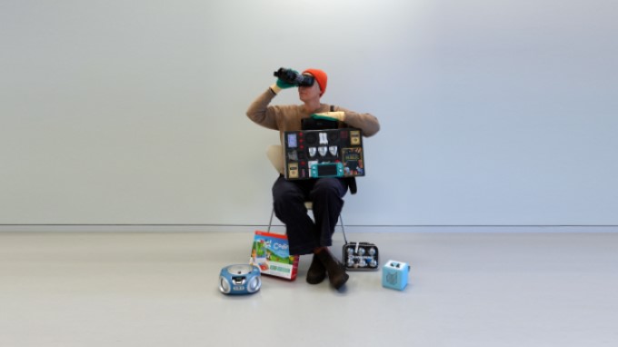 Symbolbild Bibliothek der Dinge. Eine Person sitzt auf einem Hocker und ist umgeben mit verschiedenen geräten und Dingen die in der Bibliothek ausgeliehen werden können