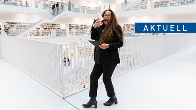 Eine Frau steht mit einem Megafon im Galeriesaal der Stadtbibliothek am Mailänder Platz. Auf einem blauen Balken steht in weißer Schrift AKTUELL